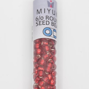 May include: A clear tube filled with small, round, red seed beads. The tube has a white label with the text "MIYU 6/0 ROUND SEED BE" and a blue and white graphic. The beads appear to be made of glass or a similar material.
