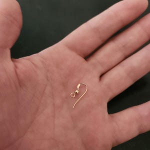 Puede incluir: Un solo gancho de pendiente de color dorado con una peque&ntilde;a bola y un alambre curvado. El gancho del pendiente se sostiene en la mano abierta de una persona, contra un fondo oscuro. El gancho est&aacute; dise&ntilde;ado para orejas perforadas.