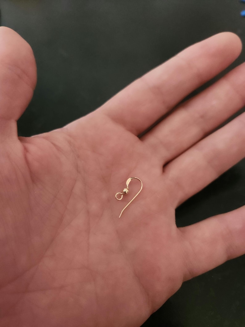 Puede incluir: Un solo gancho de pendiente de color dorado con una peque&ntilde;a bola y un alambre curvado. El gancho del pendiente se sostiene en la mano abierta de una persona, contra un fondo oscuro. El gancho est&aacute; dise&ntilde;ado para orejas perforadas.