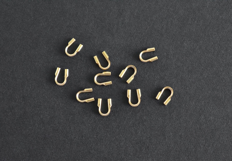 May include: A collection of small, gold-colored U-shaped jewelry findings scattered on a dark gray surface. Each finding has a curved body with two parallel, open ends, designed for connecting jewelry components.