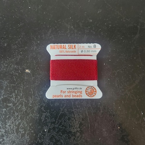 May include: A spool of red natural silk thread for stringing pearls and beads. The label reads "NATURAL SILK" and "100% Naturseide". The thread is 2 meters long and 0.80 mm in diameter. The brand name is Griffin.