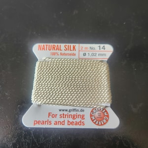 May include: A spool of natural silk thread for stringing pearls and beads. The spool is labeled "NATURAL SILK 2 m No. 14 100% Naturseide Ø 1,02 mm" and "For stringing pearls and beads www.griffin.de".