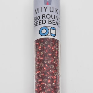 May include: A clear tube filled with small, round, red seed beads. The tube has a label with the text "MIYUKI 8/0 ROUND SEED BEAD" in blue and white. The beads are a deep red color.