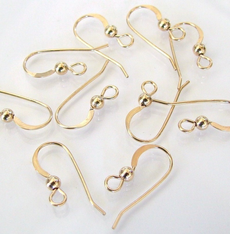 Puede incluir: Una colecci&oacute;n de ganchos para pendientes de color dorado. Cada gancho presenta una peque&ntilde;a cuenta redonda y un dise&ntilde;o curvo para asegurar los pendientes. Los ganchos se muestran sobre un fondo blanco, destacando su delicada artesan&iacute;a.