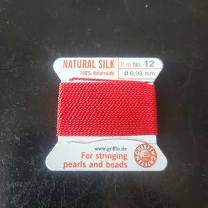 May include: A spool of red natural silk thread, labeled "NATURAL SILK" and "100% Naturseide." The label also indicates the thread is 2 meters long, with a diameter of 0.98 mm, and is for stringing pearls and beads.