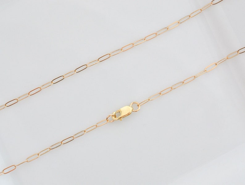 May include: A delicate gold-colored chain necklace with an elongated oval link design. The necklace features a lobster clasp closure. The chain is displayed against a white background.