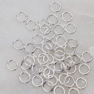 May include: A collection of small, shiny, silver-colored jump rings scattered across a light-colored surface. These circular rings are open and ready for use in jewelry making or other craft projects. The rings are uniform in size and shape.