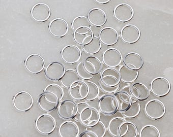 May include: A collection of small, shiny, silver-colored jump rings scattered across a light-colored surface. These circular rings are open and ready for use in jewelry making or other craft projects. The rings are uniform in size and shape.