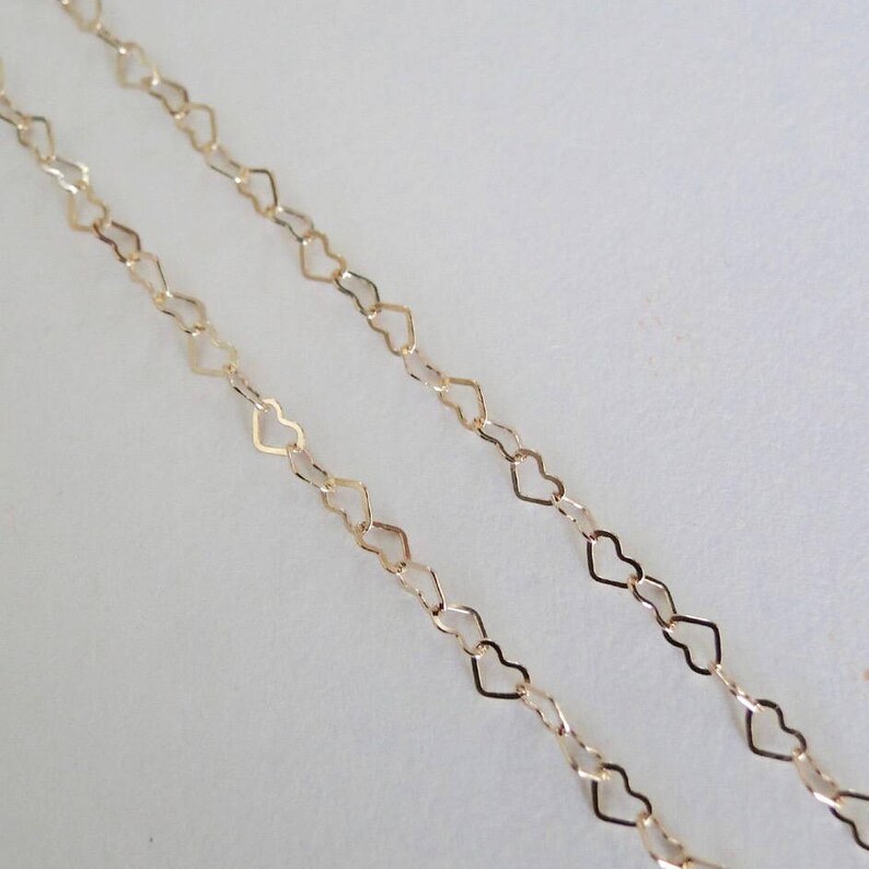 May include: Two strands of gold heart-shaped chain, one on top of the other, against a white background.