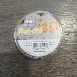 Op de afbeelding: Een spoel met 49-strengs nylon-gecoate 24 karaats vergulde roestvrijstalen microdraad, gemarkeerd als "flex-rite" door Bead Smith. De spoel is wit met gouden letters en een barcode. Hij is 10 voet (3,06 meter) lang en heeft een breeksterkte van 12,8 lb. Het productnummer is #FL49X018GP10.