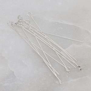 May include: A collection of silver-colored head pins with looped ends, scattered on a light gray surface. The pins are thin and straight, with a small loop at one end, and are likely used for jewelry making or crafting.