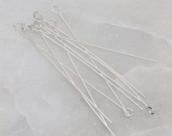 May include: A collection of silver-colored head pins with looped ends, scattered on a light gray surface. The pins are thin and straight, with a small loop at one end, and are likely used for jewelry making or crafting.