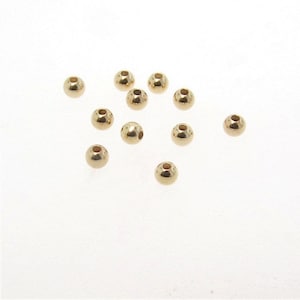 May include: A collection of small, shiny, gold-colored metal beads. Each bead is spherical with a small hole running through the center. The beads are scattered on a white surface, reflecting light.