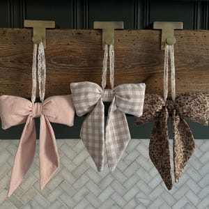 Puede incluir: Tres lazos decorativos colgando de ganchos de latón sobre una tabla de madera. Los lazos son de color rosa, cuadros vichy beige y estampados florales marrones. Cada lazo está suspendido por una delicada cinta de encaje, creando una estética encantadora y rústica.