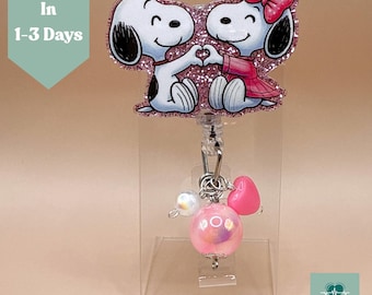 May include: A badge reel featuring a glittery Snoopy and Woodstock design, with the characters forming a heart shape. The reel includes pink heart and bead charms. The text "Ships In 1-3 Days" is visible.