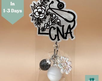 May include: A CNA badge reel with a black and white floral design, a stethoscope, and the letters CNA. The badge reel has a clear plastic case and decorative beads. The text "Ships In 1-3 Days" is visible.