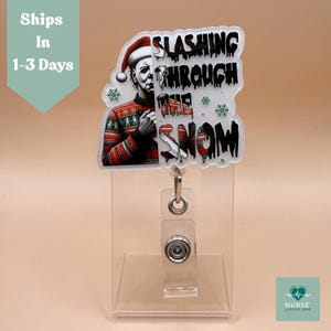 May include: A badge reel featuring a graphic of Michael Myers in a Santa hat and a Christmas sweater. The text reads "Slashing Through The Snow." The reel has a clear plastic holder and a metal clip.