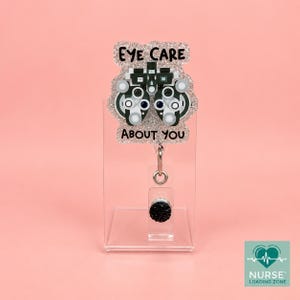 May include: A glittery badge reel featuring a phoropter illustration with the text EYE CARE ABOUT YOU. The badge reel has a silver metal clip and a small black circular accent, attached to a clear acrylic stand for display purposes.
