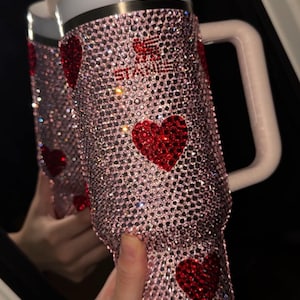 May include: A pink, bedazzled tumbler with red heart accents. The tumbler has a white lid and handle, and a straw. The text "STAY" is visible in red rhinestones. The tumbler is covered in sparkling rhinestones.