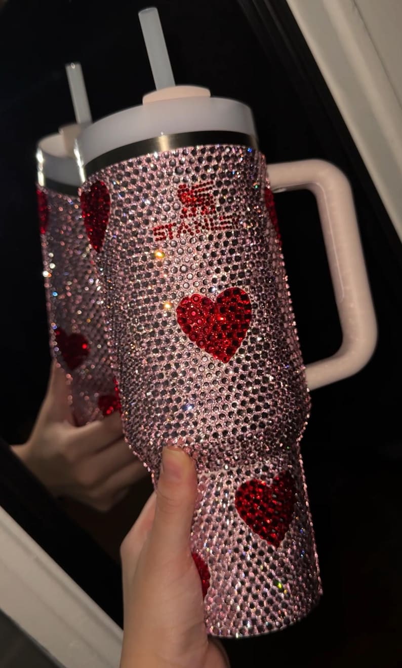 May include: A pink, bedazzled tumbler with red heart accents. The tumbler has a white lid and handle, and a straw. The text "STAY" is visible in red rhinestones. The tumbler is covered in sparkling rhinestones.