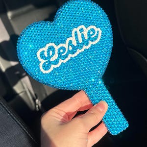 May include: A blue heart-shaped hand mirror covered in rhinestones with the word "Jessie" written in white on the front.