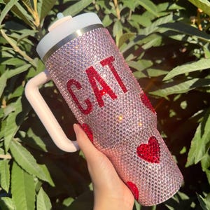 May include: Pink bedazzled tumbler with the word "CAT" in red rhinestones and red heart accents. The tumbler has a white lid, a light pink handle, and a straw. The tumbler is covered in small, sparkling rhinestones.