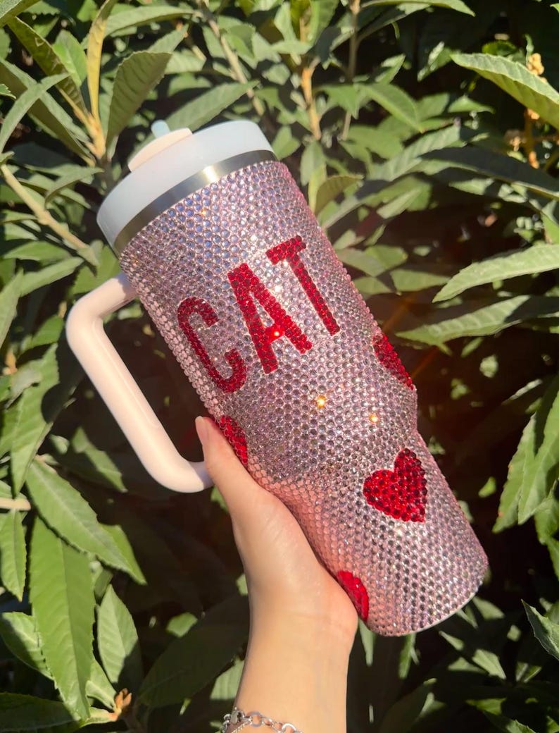 May include: Pink bedazzled tumbler with the word "CAT" in red rhinestones and red heart accents. The tumbler has a white lid, a light pink handle, and a straw. The tumbler is covered in small, sparkling rhinestones.