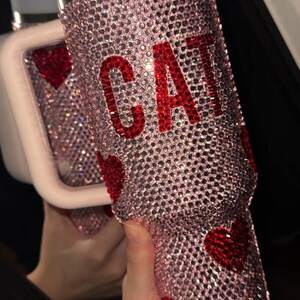 May include: A pink, bedazzled tumbler with the word "CAT" in red rhinestones. The tumbler is covered in small, sparkling crystals and features red heart accents. The lid is white with a silver band.