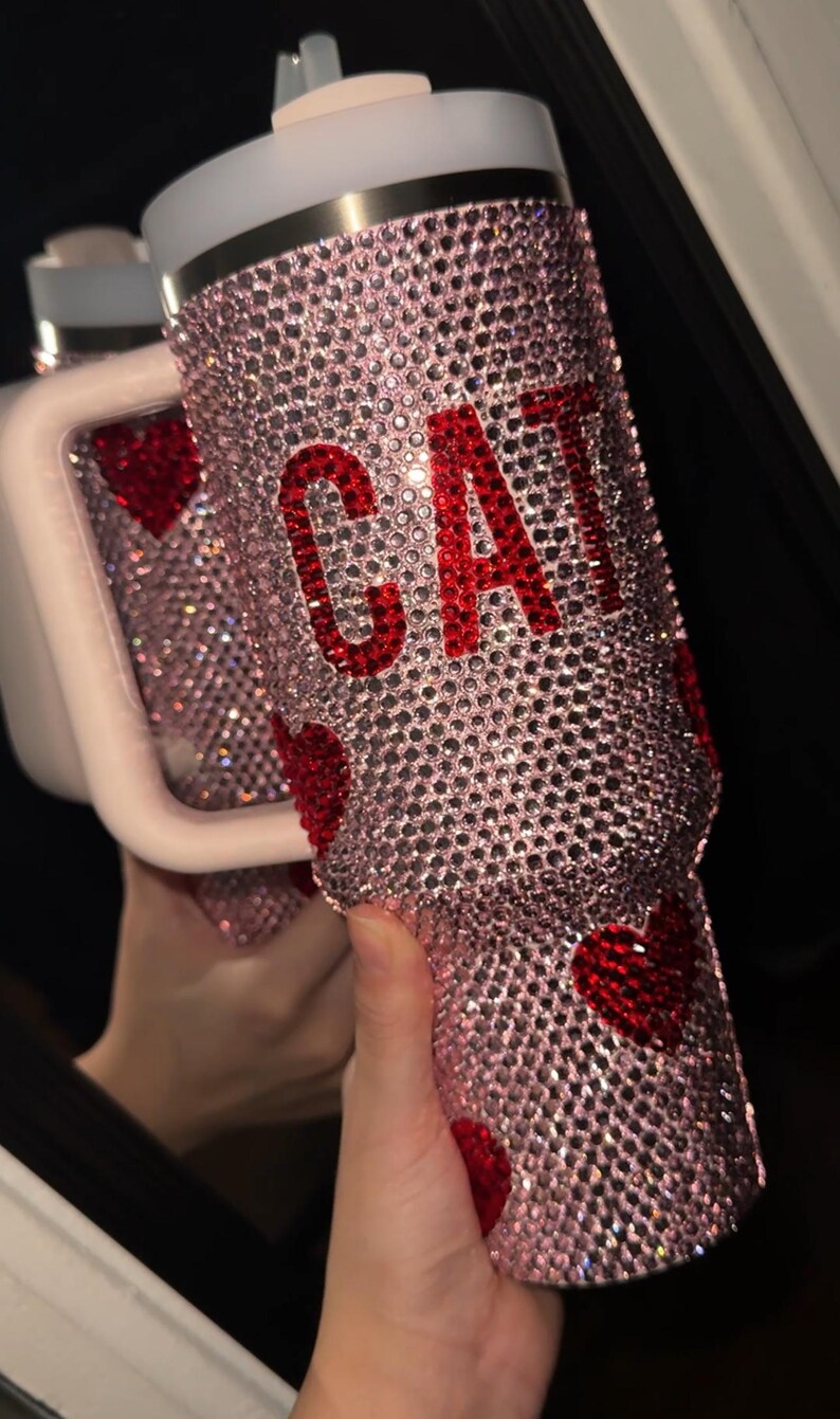May include: A pink, bedazzled tumbler with the word "CAT" in red rhinestones. The tumbler is covered in small, sparkling crystals and features red heart accents. The lid is white with a silver band.