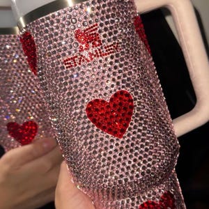 May include: Pink rhinestone-covered tumbler with red heart accents. The tumbler has a white handle, a white lid, and a clear straw. The design includes the text "Stay Sweet". The tumbler is designed to hold approximately 30 ounces.