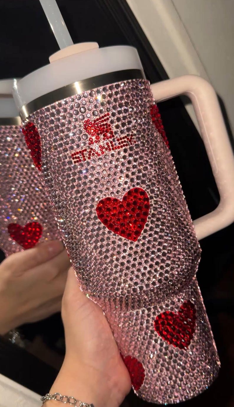May include: Pink rhinestone-covered tumbler with red heart accents. The tumbler has a white handle, a white lid, and a clear straw. The design includes the text "Stay Sweet". The tumbler is designed to hold approximately 30 ounces.