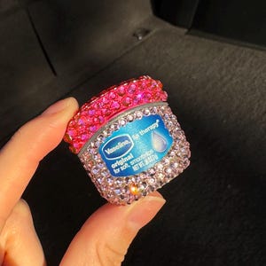 May include: A small pink and clear container of Vaseline lip therapy with rhinestones covering the container. The label reads "Vaseline lip therapy original for soft, smooth lips. Net Wt. 0.15 oz (4.25 g)"