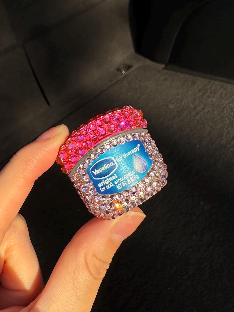 May include: A small pink and clear container of Vaseline lip therapy with rhinestones covering the container. The label reads "Vaseline lip therapy original for soft, smooth lips. Net Wt. 0.15 oz (4.25 g)"