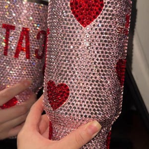 May include: A pink tumbler covered in sparkling rhinestones, with red heart accents. The tumbler has a white lid and a straw. The word "CAT" is visible in red rhinestones on a second tumbler in the background.
