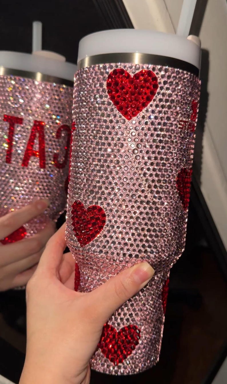 May include: A pink tumbler covered in sparkling rhinestones, with red heart accents. The tumbler has a white lid and a straw. The word "CAT" is visible in red rhinestones on a second tumbler in the background.