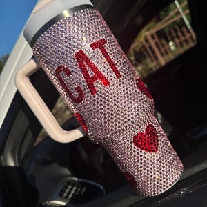 May include: A pink bedazzled tumbler with the word "CAT" in red rhinestones. The tumbler has a handle and a lid. Red heart-shaped rhinestones are also on the tumbler.