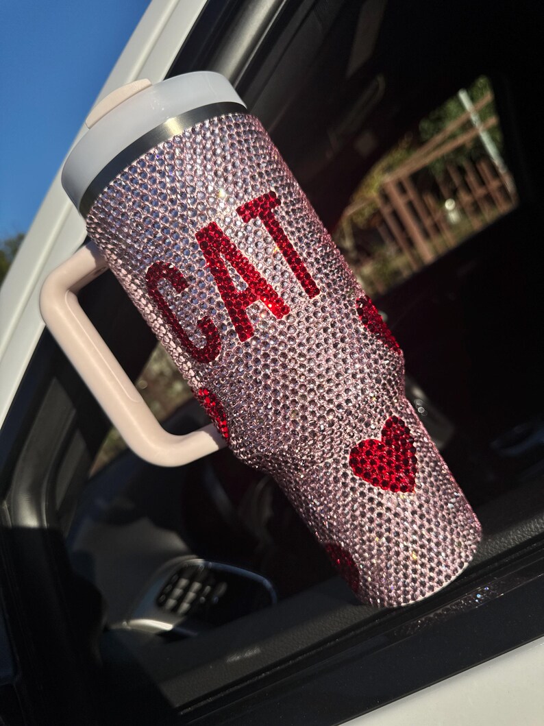 May include: A pink bedazzled tumbler with the word "CAT" in red rhinestones. The tumbler has a handle and a lid. Red heart-shaped rhinestones are also on the tumbler.
