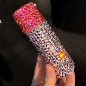 May include: A pink and clear bottle covered in rhinestones. The bottle is decorated with small, iridescent rhinestones.