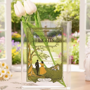 Op de afbeelding: Een heldere acrylvaas met een decoratief paneel met de boekomslag van "Wuthering Heights" van Emily Brontë. Het paneel toont een stel en een huis onder een boom. Twee witte tulpen staan in de vaas.