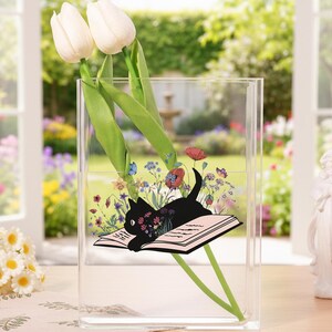 Può includere: Un vaso rettangolare trasparente con un'illustrazione di un gatto nero sdraiato su un libro in mezzo a fiori colorati. Due tulipani bianchi sono posti all'interno del vaso. Lo sfondo mostra un giardino con una fontana e una statua di angelo bianco.