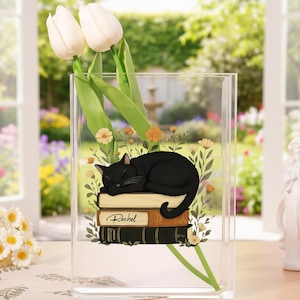 Op de afbeelding: Een heldere acrylvaas met een illustratie van een zwarte kat die op boeken slaapt. De vaas bevat witte tulpen en bloemdecoraties. Rechts is een wit engelenbeeld te zien. De achtergrond toont een tuin.