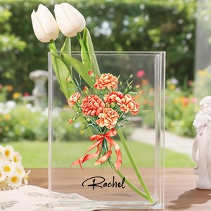 May include: A clear acrylic vase holding a bouquet of orange carnations and white tulips. The carnations are tied with a red ribbon. The name "Rachel" is printed in black script on the vase. A small white angel statue is to the right.