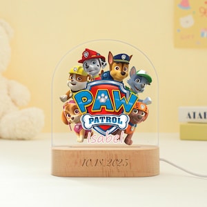 Może przedstawiać: Lampka nocna Paw Patrol z logo i postaciami z Paw Patrol. Światło ma drewnianą podstawę z wygrawerowaną datą 10.18.2025. Wyświetlana jest również nazwa "Isabel".