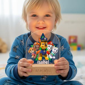 Może przedstawiać: Lampka nocna z motywem Psiego Patrolu z postaciami Chase'a, Marshalla, Skye, Rubble'a i Everest. Drewniana podstawa z wygrawerowanym imieniem "Isabel" i datą "10.18.2025". Postacie na niebieskiej gwieździe. Świecące światło.