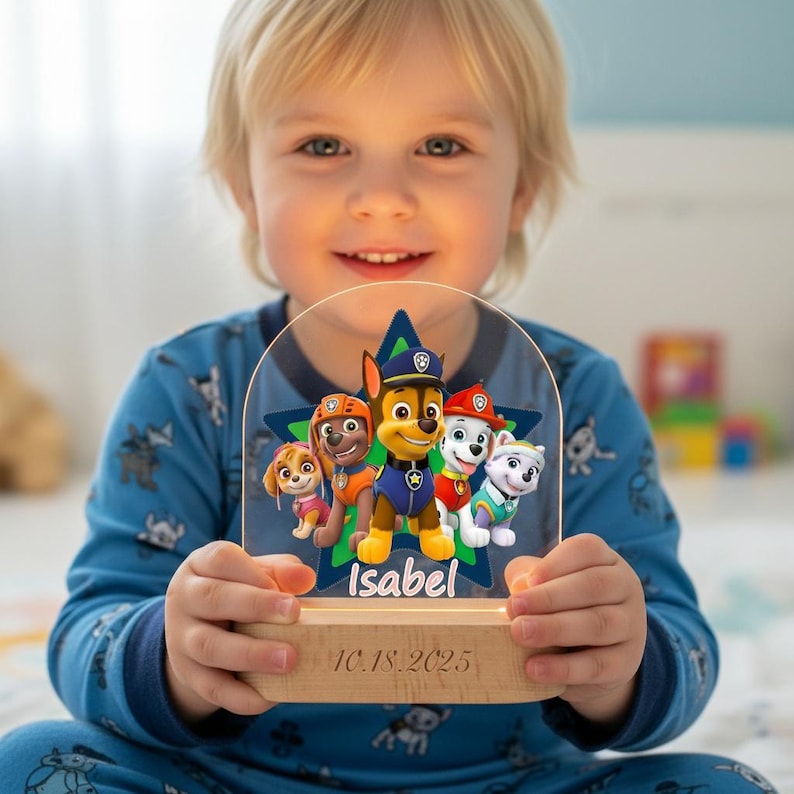 May include: A Paw Patrol night light featuring the characters Chase, Marshall, Skye, Rubble, and Everest. The light has a wooden base with the name "Isabel" and the date "10.18.2025" engraved. The characters are set against a blue star.