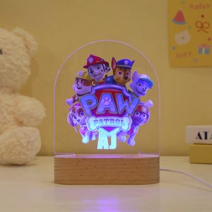 May include: A Paw Patrol night light with a wooden base. The clear acrylic panel features the Paw Patrol characters and the Paw Patrol logo. The light is powered by a cord and has the initials "AJ" at the bottom.