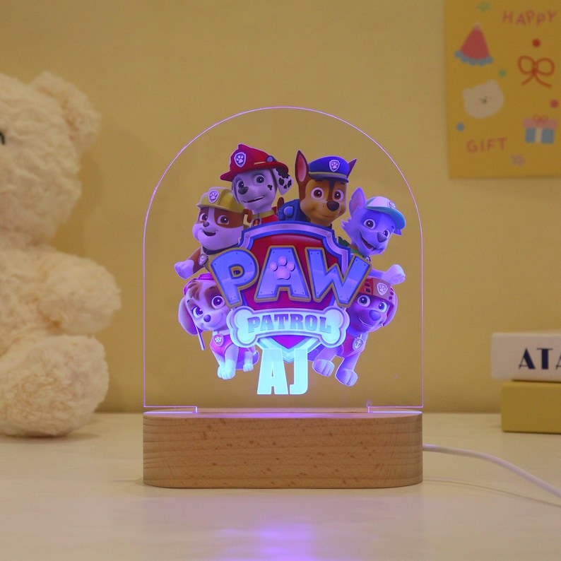 May include: A Paw Patrol night light with a wooden base. The clear acrylic panel features the Paw Patrol characters and the Paw Patrol logo. The light is powered by a cord and has the initials "AJ" at the bottom.