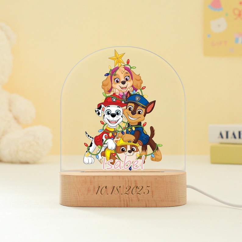 May include: A decorative night light featuring a Paw Patrol design. The light has a wooden base with the date "10.18.2025" and the name "Isabel" engraved. The light has a clear, arched top with the Paw Patrol characters.