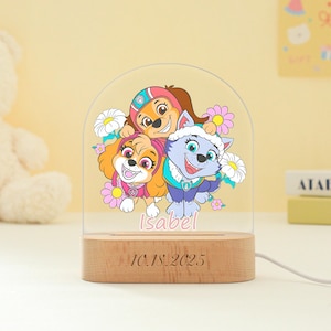 May include: A decorative night light featuring a colorful illustration of three cartoon puppies surrounded by flowers. The light has a wooden base with the date "10.18.2025" engraved on it, and the name "Isabel" is written in pink. A white cord extends from the base.