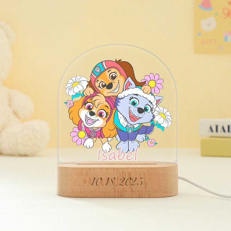May include: A decorative night light featuring a colorful illustration of three cartoon puppies surrounded by flowers. The light has a wooden base with the date "10.18.2025" engraved on it, and the name "Isabel" is written in pink. A white cord extends from the base.